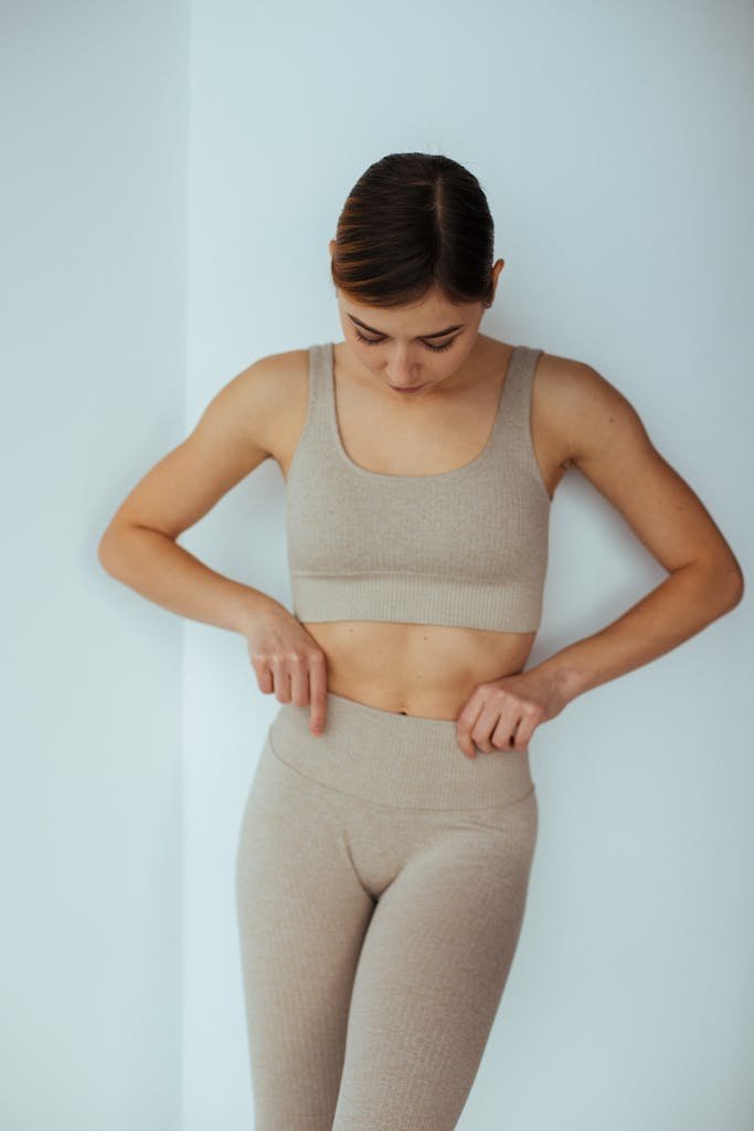 Young woman adjusting her fitted beige sportswear indoors for a modern fitness look.